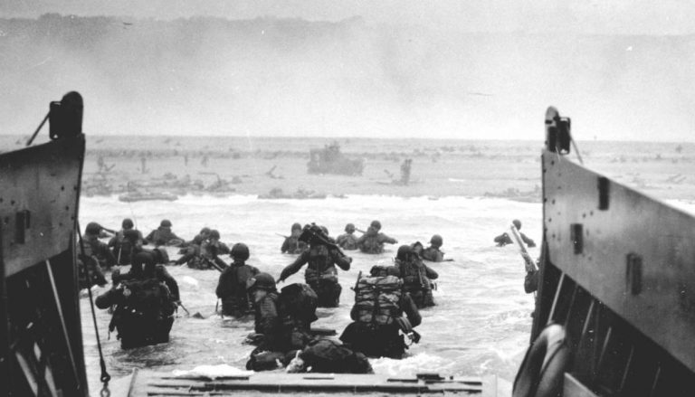 Soldiers, transitioning from veteran life, wade through water towards a mist-covered beach from a landing craft.