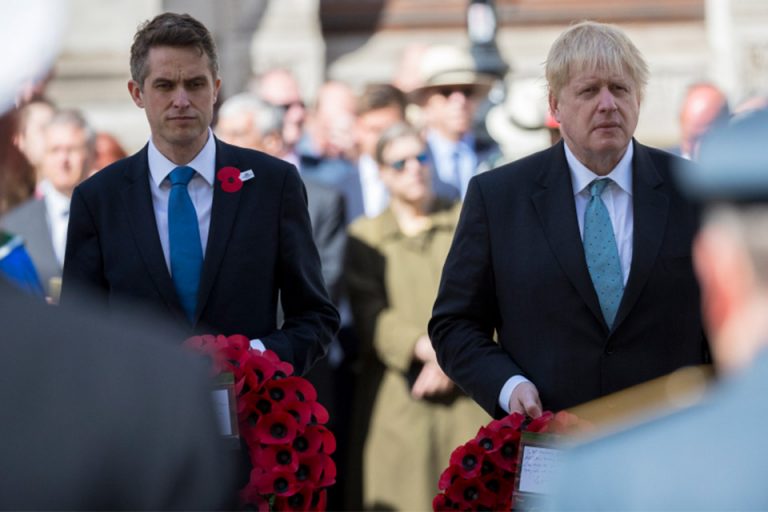 Two men in suits holding wreaths of red poppies stand solemnly at a military event, with a crowd gathered respectfully in the background.