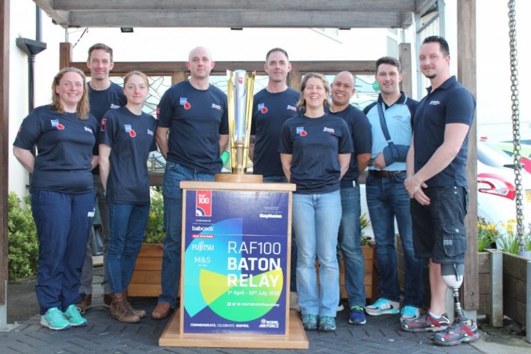 A group of people in matching shirts, including a proud Army veteran, stand around a large trophy labeled "RAF100 Baton Relay," under a wooden structure outdoors.