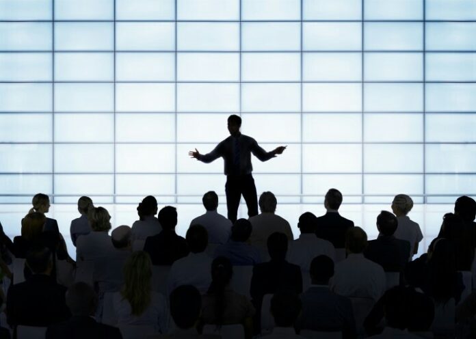 A person gives a presentation to an audience in front of a large illuminated grid screen, discussing military strategies and the challenges of resettlement after deployment.
