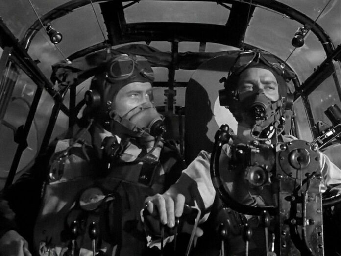 Two veteran pilots in vintage flight gear sit in a cockpit, both wearing oxygen masks and helmets, surrounded by military flight instruments and controls.
