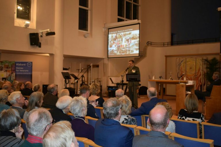 A veteran speaker addresses an audience in a large room, seamlessly sharing their journey from service to resettlement. The presentation screen displays images of navigating Civvy Street, offering inspiration and guidance to those embarking on a new path.