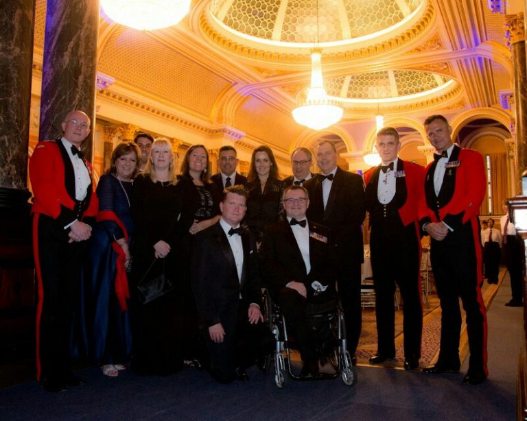 A group of veterans in formal attire, including some in red military uniforms, pose indoors under ornate lighting and architecture, celebrating their resettlement from Civvy Street.