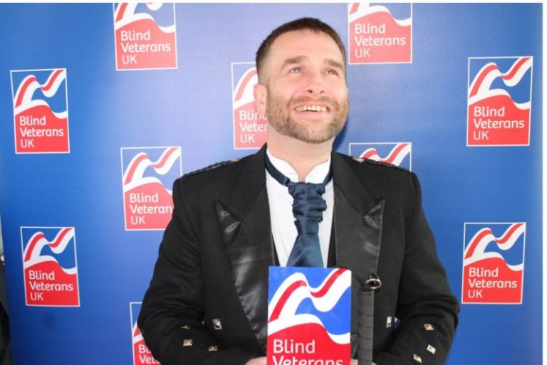 A smiling man in formal attire holds an award against a backdrop featuring "Blind Veterans UK" logos, celebrating his successful resettlement into Civvy Street.