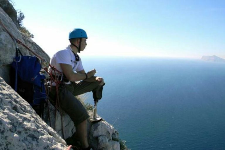 A climber with a prosthetic leg sits on a cliff edge, wearing a helmet and harness, like a pathfinder charting new routes, gazing at the vast ocean below.