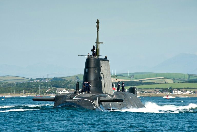 A Royal Navy submarine navigates through a body of water, with land and hills visible in the background. It is partially surfaced with crew members standing on top, embodying steadfast dedication amidst ever-changing resettlements and landscapes.
