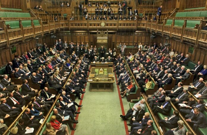 Interior view of a packed parliamentary chamber with members seated on green benches, listening attentively as a veteran shares his journey from the Royal Navy to becoming a political pathfinder.