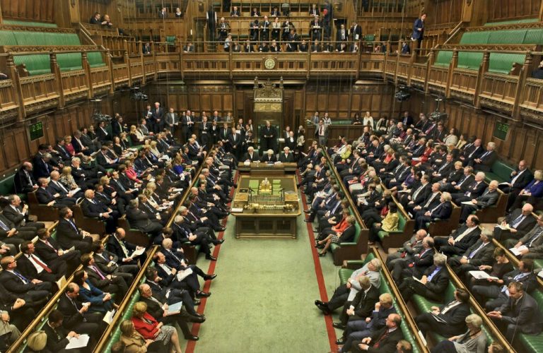 Interior view of a packed parliamentary chamber with members seated on green benches, listening attentively as a veteran shares his journey from the Royal Navy to becoming a political pathfinder.