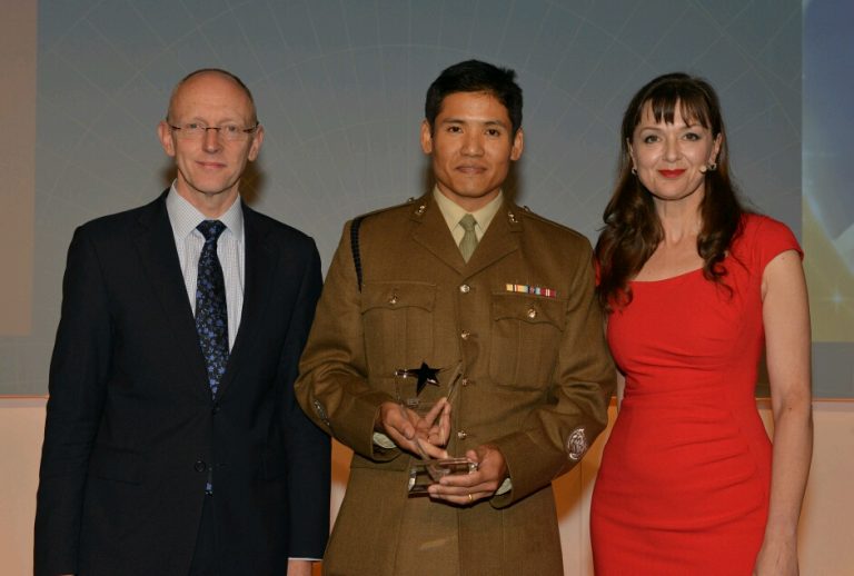 Three individuals stand together against a light background: one in a suit, another in a military uniform holding a trophy celebrating an Army achievement, and the third in a striking red dress.