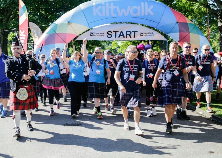 Participants wearing kilts start a charity walk under a colorful arch, inspired by veterans transitioning from Civvy Street, with a bagpiper leading them.