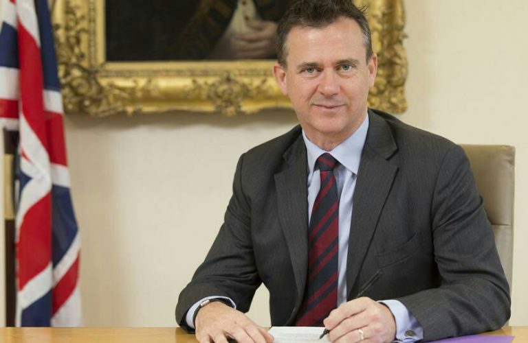 A man in a suit sits at a desk, pen poised over paper, as he contemplates his resettlement into civvy street. Behind him, a British flag and a framed portrait symbolize his past as a veteran.