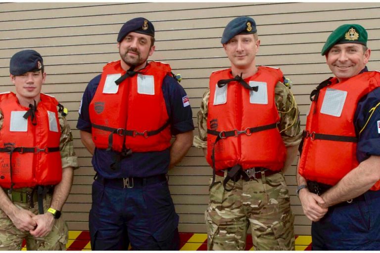 Four veterans in military uniforms and berets, wearing orange life vests, stand proudly in front of a striped wall.