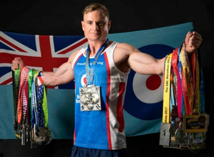 A veteran in a sports jersey proudly holds numerous medals and ribbons in front of a flag backdrop, showcasing their triumphs after resettlement into civvy street.