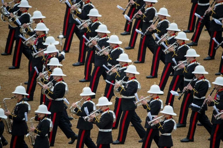Uniformed musicians in white helmets from the Royal Navy march in formation, playing brass and woodwind instruments on a parade ground.