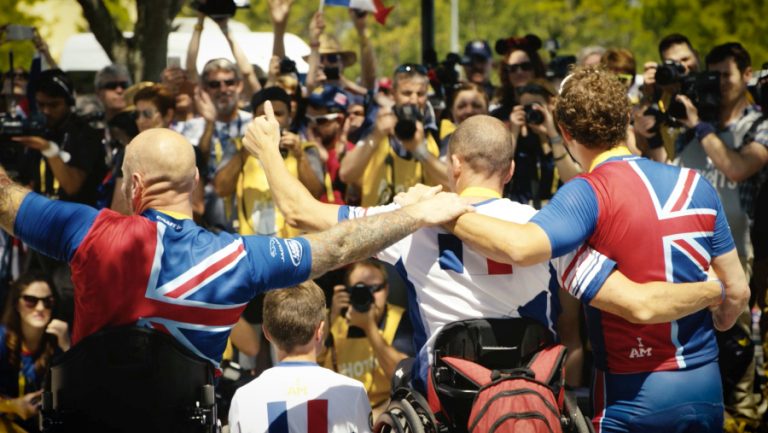 Three athletes, including a veteran in a wheelchair, celebrate facing a crowd and cameras. They wear jerseys adorned with British and French flags, embodying the spirit of camaraderie both on the field and in their journey toward resettlement.
