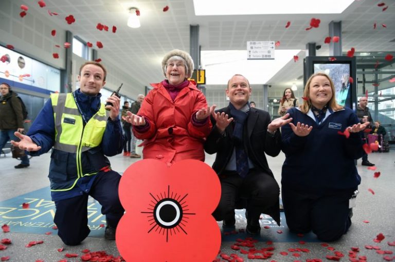Four people pose with a large poppy symbol in a station as red poppy petals fall around them, honoring veterans transitioning to Civvy Street. They appear to be participating in a commemorative event.