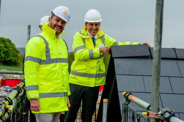 Two people in high-visibility jackets and hard hats stand on a construction site, inspecting roof tiles on a building as part of a military resettlement program.