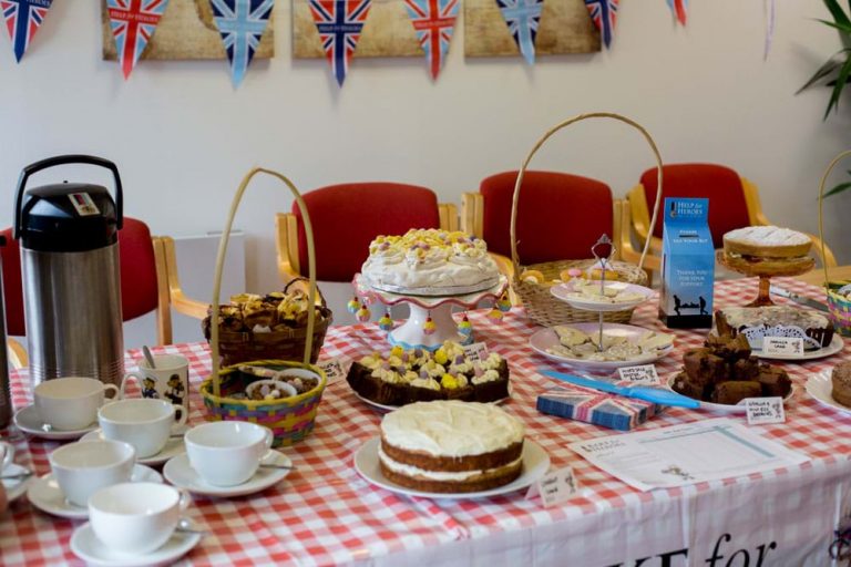 A table with various cakes, pastries, teacups, and a coffee dispenser evokes the warm embrace of civvy street. British flags and bunting decorate the background, offering a sense of nostalgia and resettlement for veterans celebrating a welcome change in life.