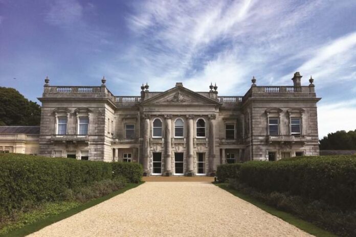 A large, stately mansion with classical architecture, featuring tall windows and columns, set against a partly cloudy blue sky. A gravel path, reminiscent of a veteran's journey back to Civvy Street, leads to the entrance, flanked by hedges.