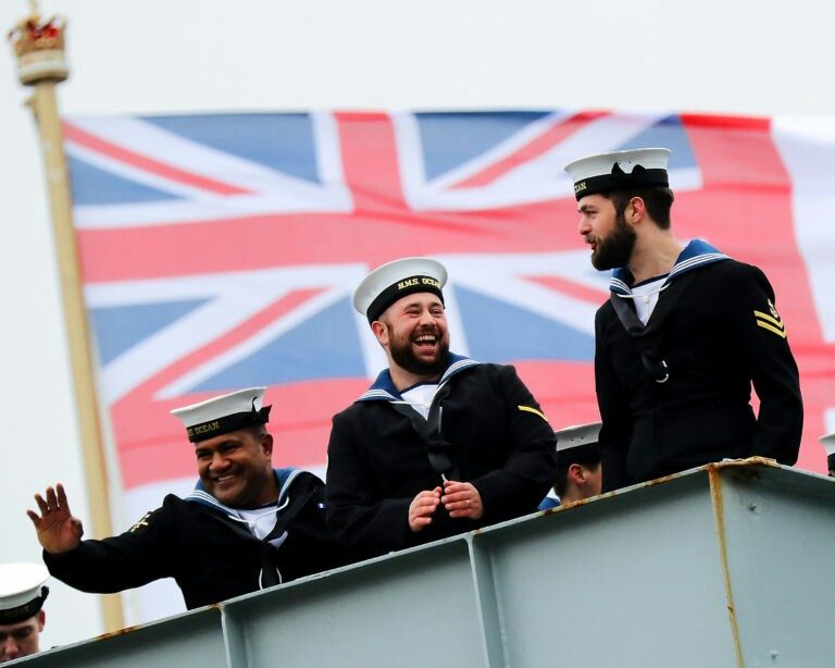 Three sailors in uniform stand proudly on a ship, smiling and waving, with the majestic British flag of the Royal Navy fluttering in the background.