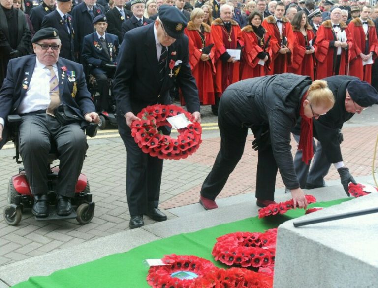 People place wreaths of red poppies on a memorial during a ceremony. A veteran in a wheelchair, adorned with medals, watches intently. The crowd, including individuals in red robes and Royal Navy uniforms, stands solemnly in the background.
