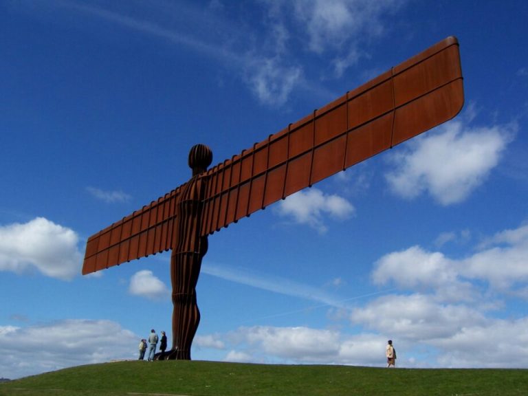On a grassy hill stands a large, rust-colored metal sculpture of an angel with wide wings, symbolizing the courage of a veteran. People gather nearby under the blue sky with scattered clouds.