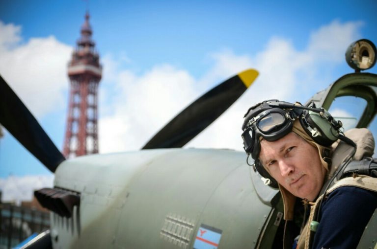 A veteran in aviation gear stands proudly beside a vintage aircraft, with a tall tower rising against the clear blue sky.
