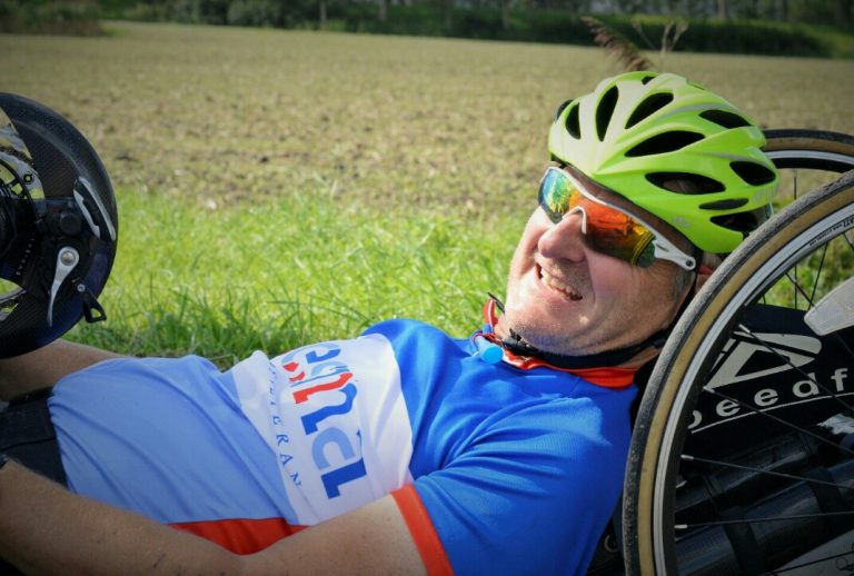 A veteran in bright cycling gear and a helmet lounges on the grass, smiling. With sunglasses shielding his eyes, he proudly rests beside his handcycle, embodying a spirit of adventure and resettlement as he embraces this new chapter after his time with the Royal Navy.