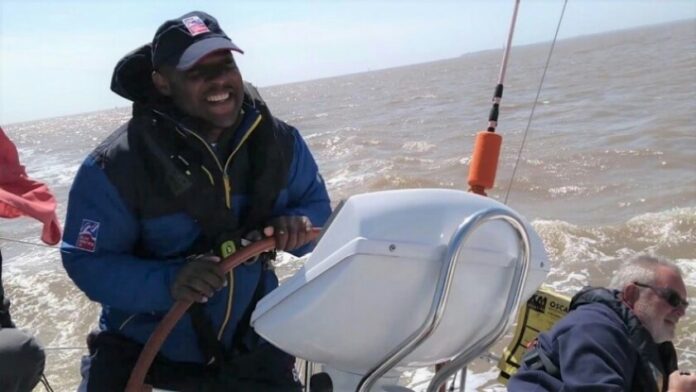 A veteran steering a sailboat on a sunny day, wearing a blue jacket and cap, smiling confidently. Another person sits nearby, partially visible as they enjoy the ocean waves in the background.