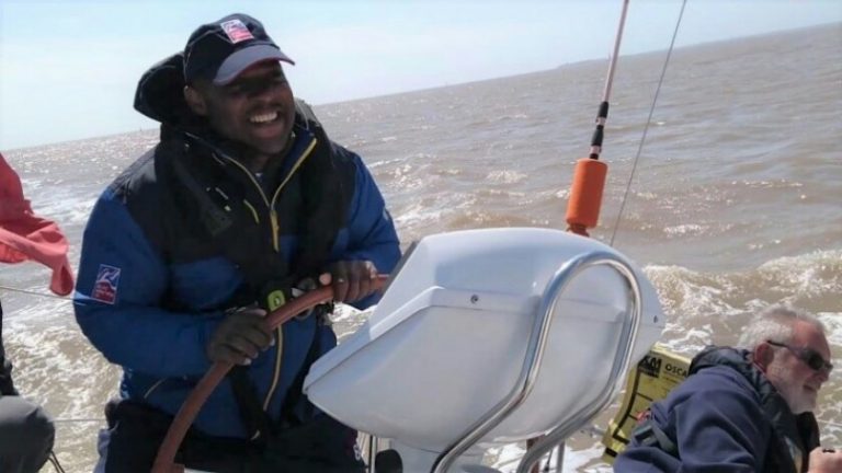 A veteran steering a sailboat on a sunny day, wearing a blue jacket and cap, smiling confidently. Another person sits nearby, partially visible as they enjoy the ocean waves in the background.