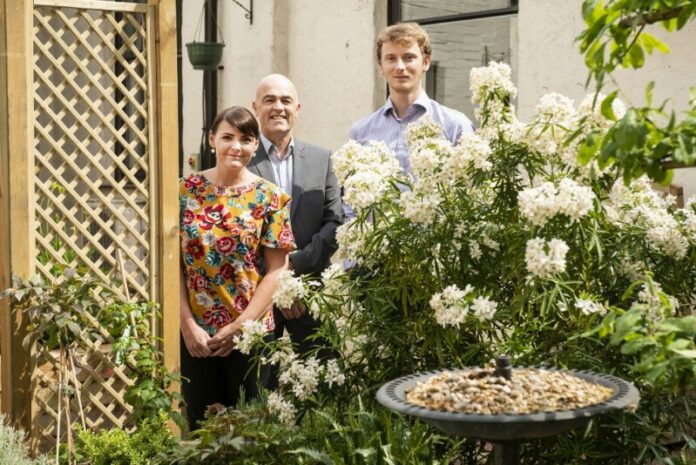 Three people stand in a garden with a trellis and flowering bushes, beside a birdbath filled with pebbles—a serene backdrop for the veteran pathfinder's journey of resettlement.
