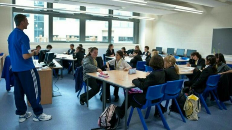 A teacher stands at the front of a classroom, speaking to a group of students seated at two long tables, reminiscent of the discipline found in a Royal Navy training session, with computers in the background.
