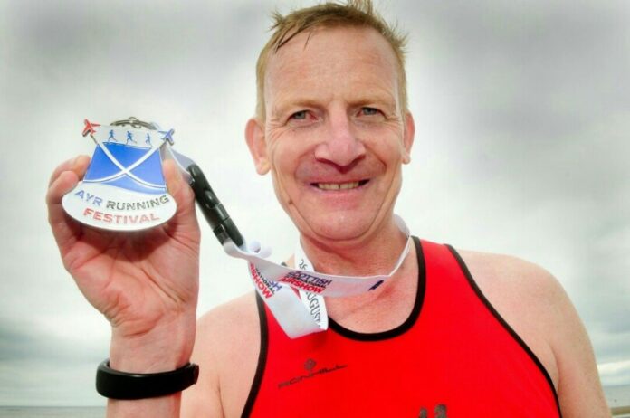 A man in a red tank top, embodying the spirit of a pathfinder, proudly holds up a medal from the Ayr Running Festival, smiling against a cloudy sky.