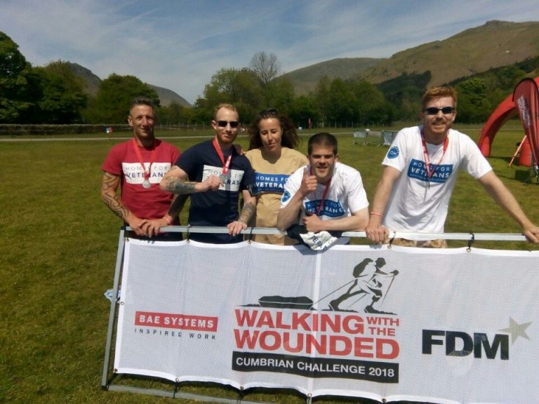 Five people stand proudly behind a banner for "Walking with the Wounded Cumbrian Challenge 2018" on a sunny day. Some sport "Homes for Veterans" shirts, showcasing their military spirit and dedication.