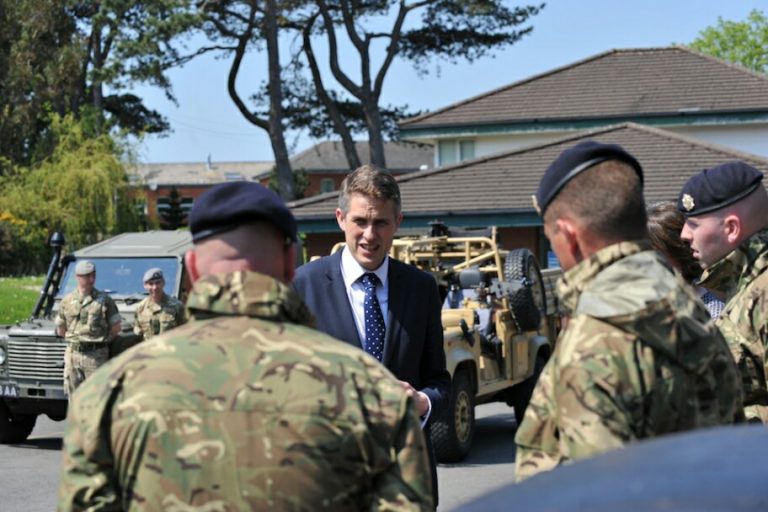 A man in a suit talks to soldiers in camouflage uniforms, discussing strategies with the seasoned precision of a veteran. A military vehicle is visible in the background, nestled among trees and a nearby house.