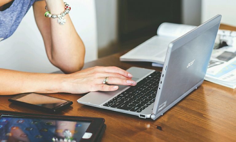 A person works on a laptop at a wooden table, their smartphone and tablet close at hand, complemented by an open magazine featuring Royal Navy stories.