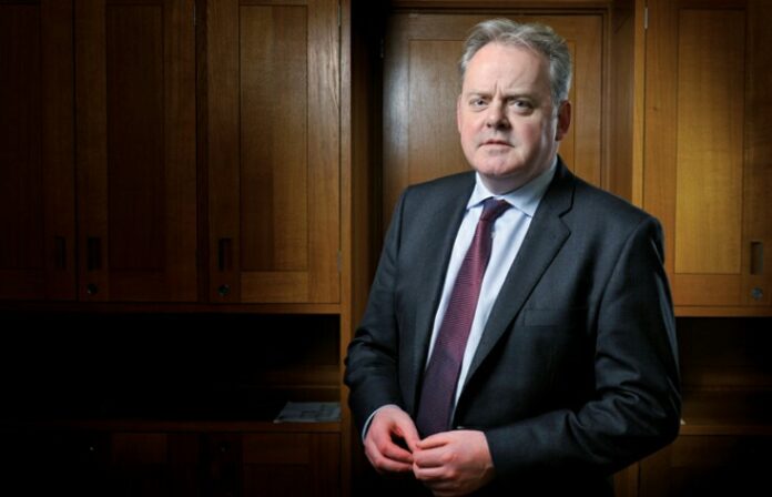 A man in a suit and tie stands proudly before wooden cabinets, exuding the composure of a seasoned Royal Navy officer.