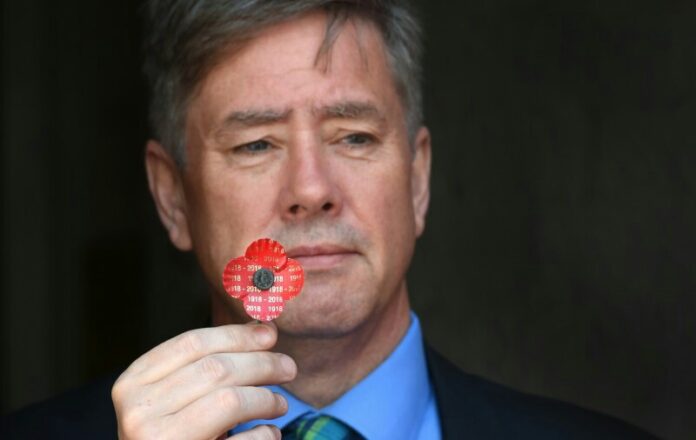 A man, possibly a veteran, holds a red remembrance poppy with dates printed on it, gazing thoughtfully. He is wearing a suit and tie, embodying the solemnity and honor associated with military service.