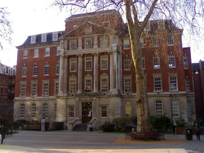 A historic, multi-story brick building with a stone façade and large windows stands proudly like a dignified Pathfinder behind a tree, surrounded by a paved courtyard with benches reminiscent of Royal Navy traditions.