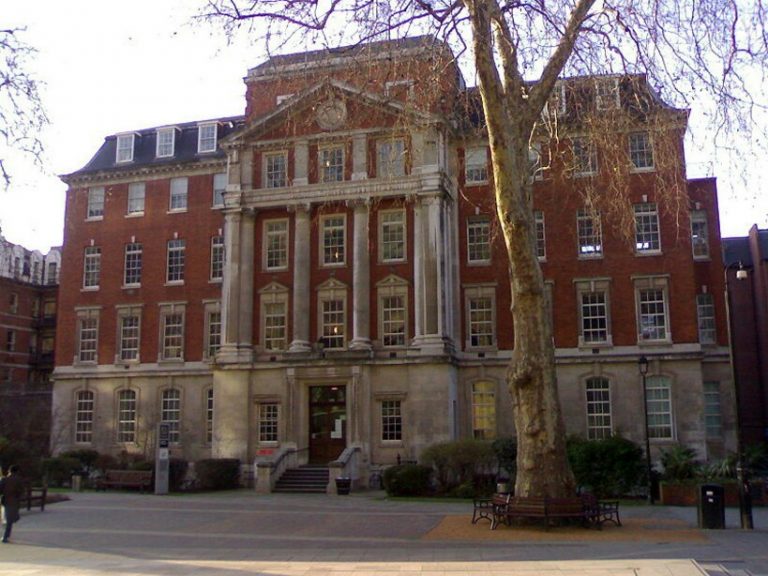 A historic, multi-story brick building with a stone façade and large windows stands proudly like a dignified Pathfinder behind a tree, surrounded by a paved courtyard with benches reminiscent of Royal Navy traditions.