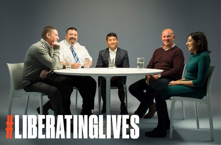 Five people, including a veteran, are sitting around a white table, smiling. The text "#LIBERATINGLIVES" is displayed prominently in the foreground.