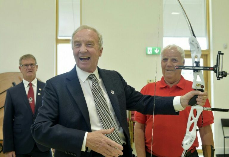 Three men stand indoors; the man in the center, a veteran, holds a white archery bow. They are wearing formal and casual attire in a light room.
