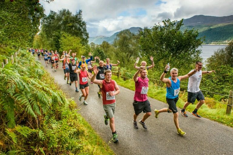 A group of runners, including a veteran, wave at the camera as they race along a scenic, tree-lined path near a shimmering body of water.