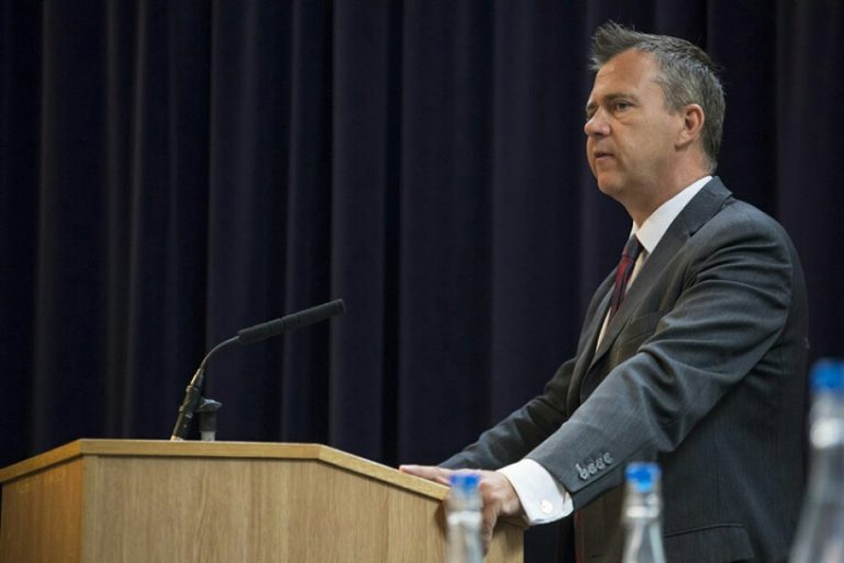 A veteran in a suit speaks at a podium with a microphone in front of a dark curtain.