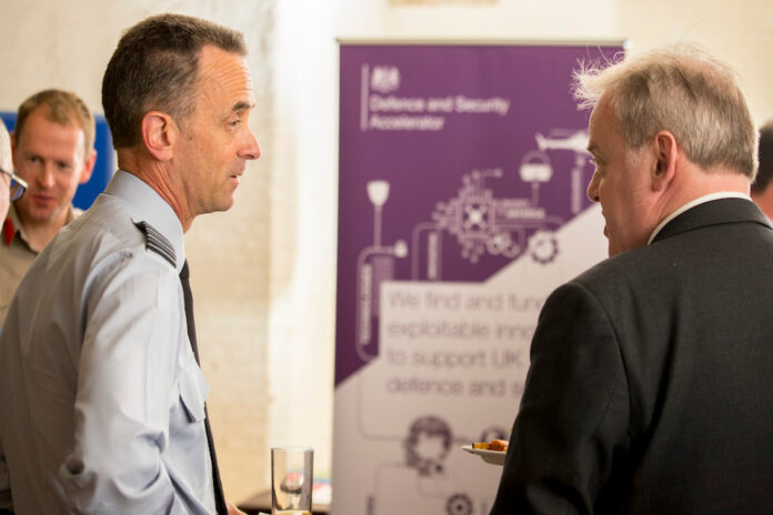 Two men engage in conversation at an event beside a banner about defense innovation. One man, holding a plate of food, discusses the Royal Navy's pathfinder initiatives and opportunities for resettlement after service.