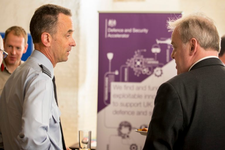 Two men engage in conversation at an event beside a banner about defense innovation. One man, holding a plate of food, discusses the Royal Navy's pathfinder initiatives and opportunities for resettlement after service.