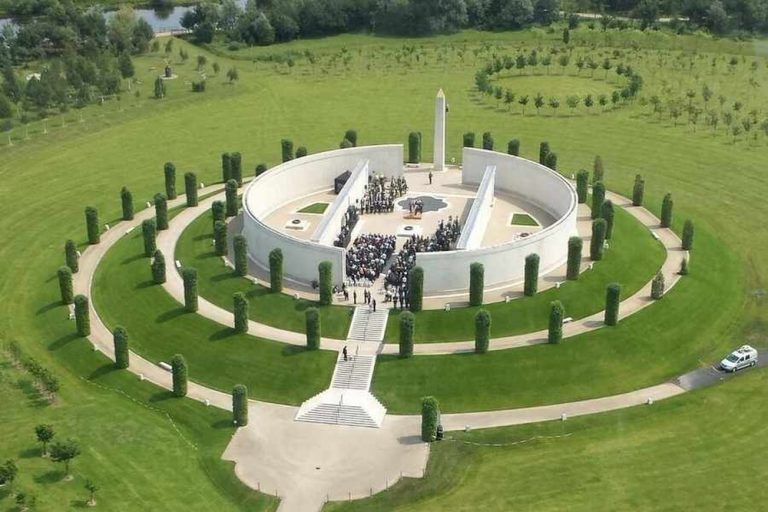 Aerial view of a circular military memorial surrounded by paths and trees, with a crowd gathered inside.