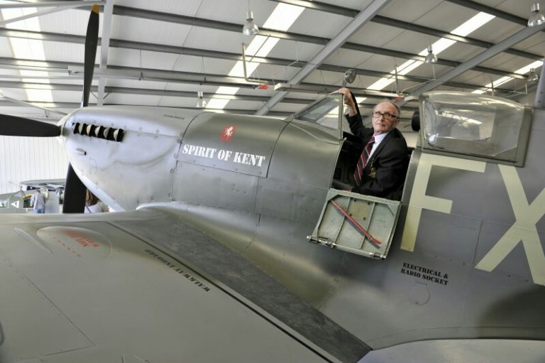 An elderly man sits in the cockpit of a vintage aircraft named "Spirit of Kent," once a pathfinder for the Royal Navy, inside a hangar.