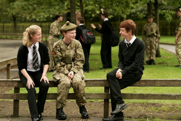 Three students sit on a bench. One wears a school uniform, another in military camouflage attire resembling an aspiring army cadet, and the last dressed in casual black clothing. Other students in uniforms fill the background.