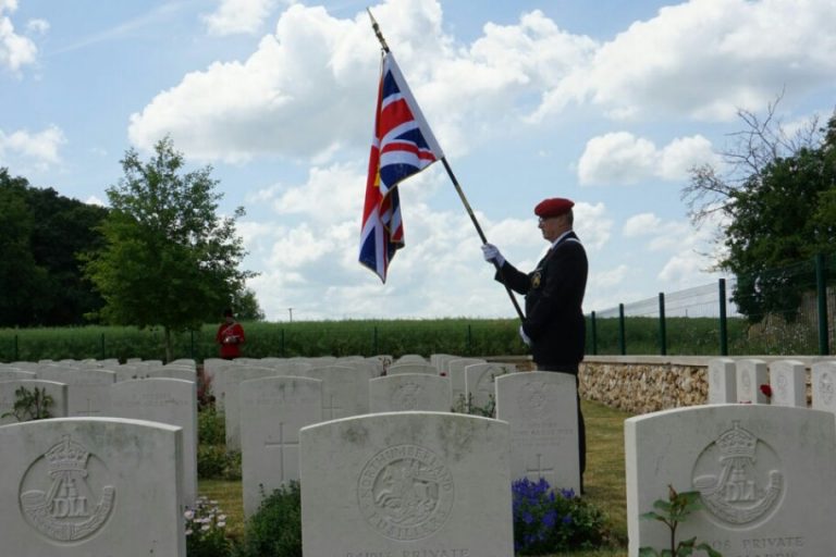A veteran in a military uniform stands solemnly in a cemetery, clutching a British flag. Headstones line the foreground, while trees sway gently under a cloudy sky, marking the pathfinder's tribute.
